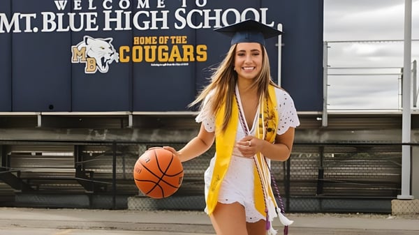 Eine Absolventin der Mount Blue High School steht in Talarm und mit Basketball vor einem Schulzeichen.