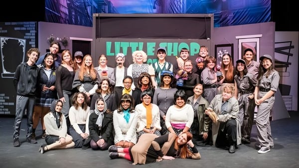 Eine Gruppe von Schülerinnen der Mount De Sales Academy posiert gemeinsam vor einem bunten urbanen Hintergrund mit dem Schriftzug Little Shop.