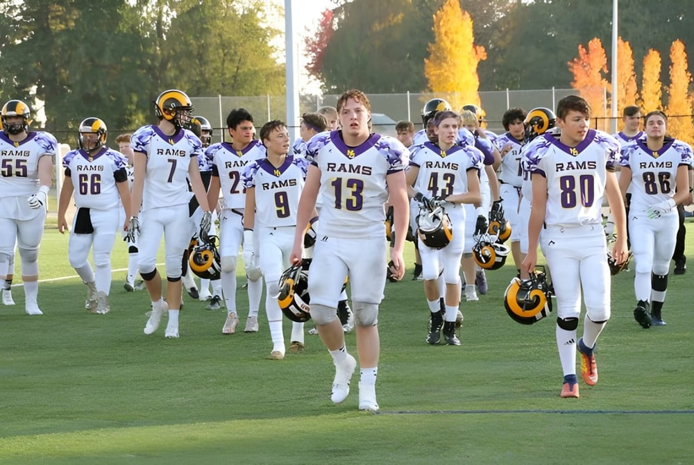 Eine Gruppe von Footballspielern in weißen Rams-Uniformen läuft auf dem Spielfeld der Mount Douglas Secondary School umgeben von herbstlichen Bäumen.