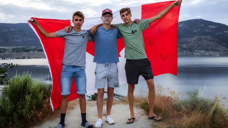 Drei Schüler der Mount Elizabeth Middle/Secondary School halten eine große rote Fahne vor einem See und Berglandschaft.