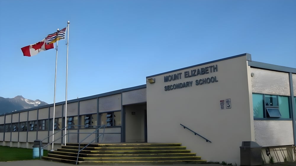 Das moderne Schulgebäude der Mount Elizabeth Middle/Secondary School mit kanadischer und amerikanischer Flagge unter klarem Himmel.