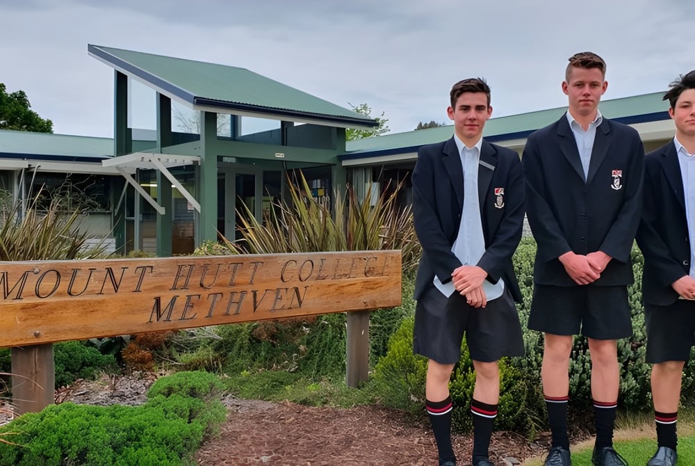 Das Holzschild von Mount Hutt College steht im Vordergrund, im Hintergrund stehen drei Schüler vor einem Gebäude mit grünem Dach.