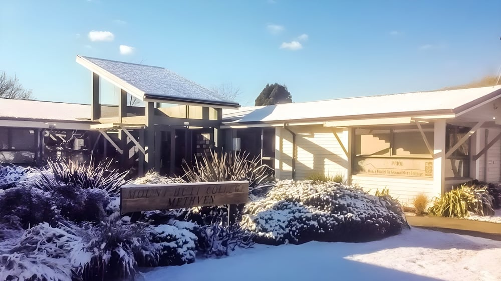 Ein schneebedecktes Holzhaus mit Solarpanelen auf dem Dach steht auf dem Campus des Mount Hutt College, umgeben von üppiger Vegetation.