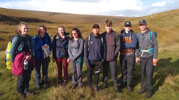 Eine Gruppe von Schülerinnen und Schülern der Mount Kelly School steht im Freien in einer hügeligen Landschaft unter bewölktem Himmel.