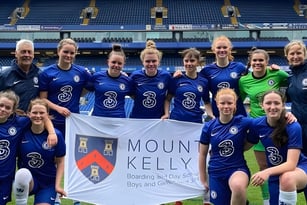 Eine Gruppe weiblicher Fußballspielerinnen der Mount Kelly School posiert gemeinsam mit einem Banner auf dem Spielfeld.