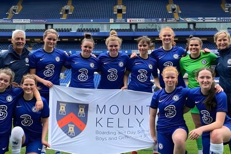 Eine Gruppe weiblicher Fußballspielerinnen der Mount Kelly School posiert gemeinsam mit einem Banner auf dem Spielfeld.