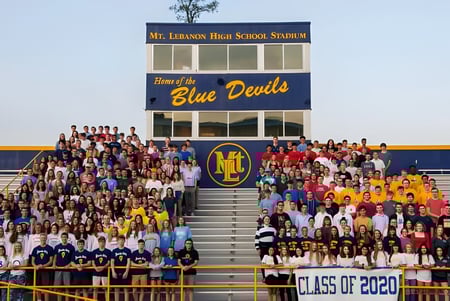 Eine Gruppe von Schülern sitzt auf den Tribünen vor dem Stadion der Mount Lebanon High School mit dem Schriftzug Home of the Blue Devils.