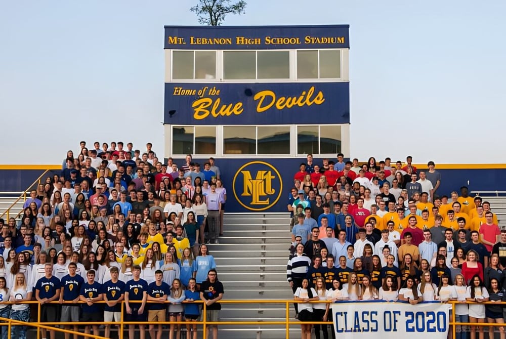Eine Gruppe von Schülern sitzt auf den Tribünen vor dem Stadion der Mount Lebanon High School mit dem Schriftzug Home of the Blue Devils.