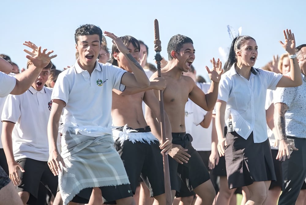 Eine Gruppe von Schülerinnen und Schülern des Mount Maunganui College steht zusammen und hebt die Hände bei strahlend blauem Himmel.