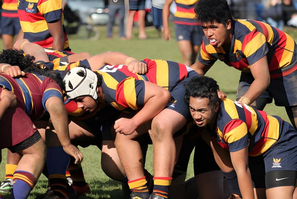 Eine Gruppe von Rugby-Spielern der Mount Roskill Grammar steht in einem Gedränge auf einem Rasenplatz mit Zuschauern im Hintergrund.