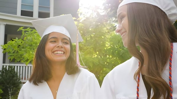Zwei Schülerinnen der Mount Saint Michael stehen in ihren Abschlussroben vor einem Haus mit Veranda und Grünpflanzen.