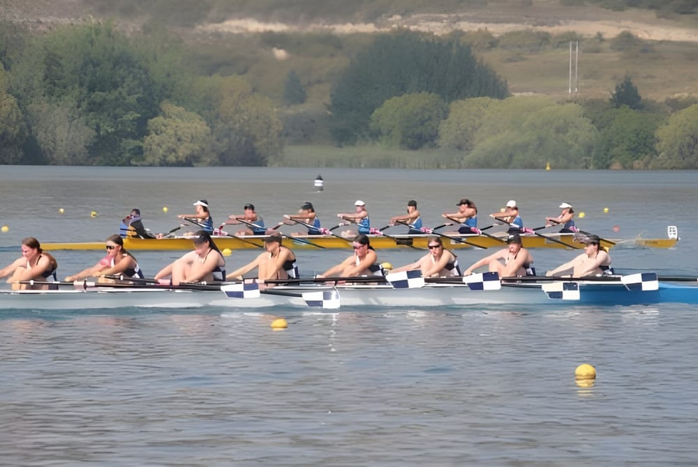 Schüler der Mountainview High School rudern in einem Langboot auf einem ruhigen See vor bewaldeten Hügeln.
