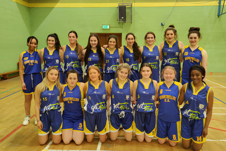 Eine Gruppe junger Basketballspielerinnen in blauen und gelben Trikots posiert in der Sporthalle der Mountmellick Community School.