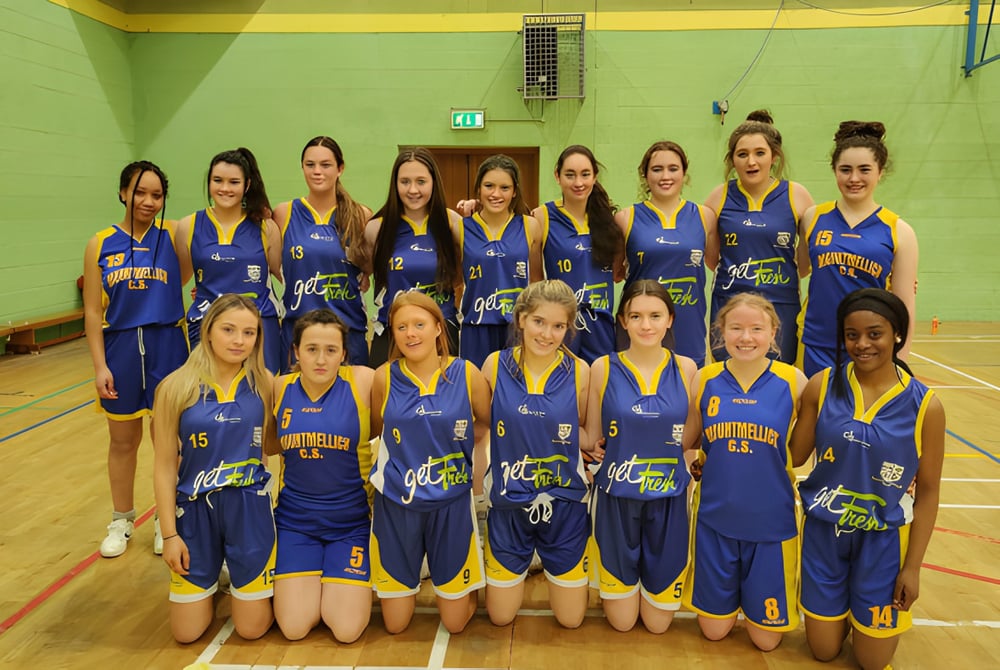 Eine Gruppe junger Basketballspielerinnen in blauen und gelben Trikots posiert in der Sporthalle der Mountmellick Community School.