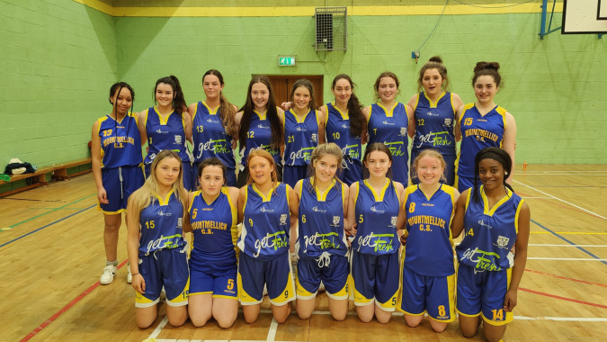 Eine Gruppe junger Basketballspielerinnen der Mountmellick Community School posiert in einer Sporthalle.