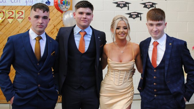 Vier junge Erwachsene in formeller Kleidung posieren vor einem bunten Abschlussdekoration auf dem Campus des Moyne College.