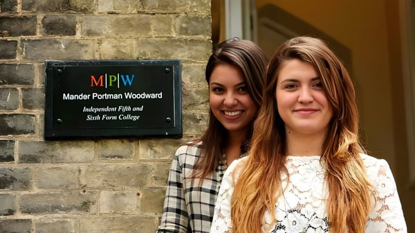 Zwei Schülerinnen mit langen dunklen Haaren stehen vor einer Backsteinwand mit dem Schild der MPW Cambridge.
