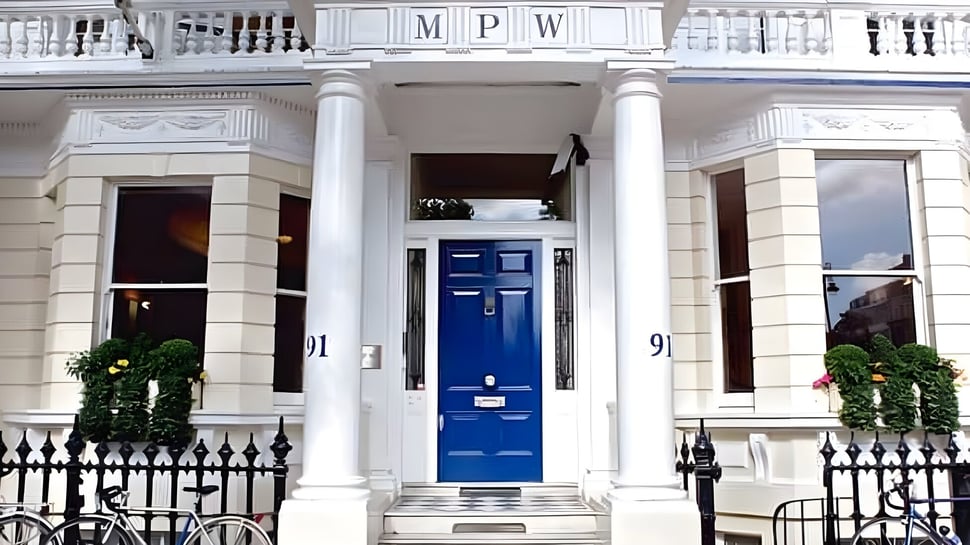 Das weiße Gebäude mit weißen Säulen und einer blauen Eingangstür auf dem Campus der MPW London ist von grünen Pflanzen umgeben.