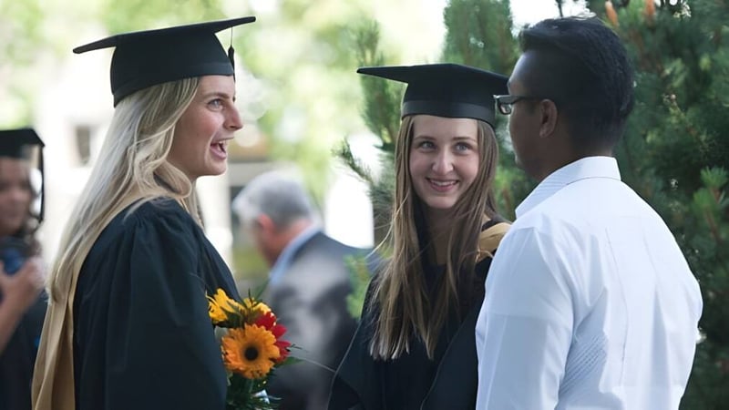 Zwei Schülerinnen der Mt. Sentinel Secondary School in Abschlussgewändern unterhalten sich mit einem Mann im Freien.