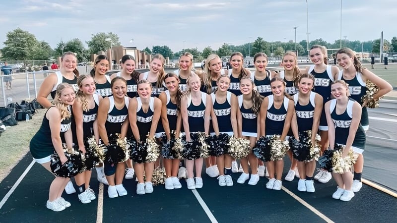 Eine Gruppe von Cheerleadern der Mt. Vernon High School posiert gemeinsam auf dem Sportplatz mit Gebäuden und Bäumen im Hintergrund.