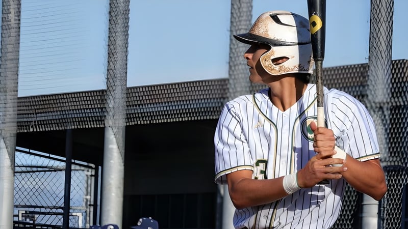 Ein Baseballspieler in weiß-blau gestreiftem Trikot steht mit einem Schläger vor einer stadionähnlichen Struktur auf dem Gelände des Murrieta Valley Unified School District.