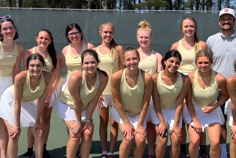 Eine Gruppe von Schülerinnen der Muskegon Catholic Central High School posiert auf dem Tennisplatz vor einem bewaldeten Hintergrund.