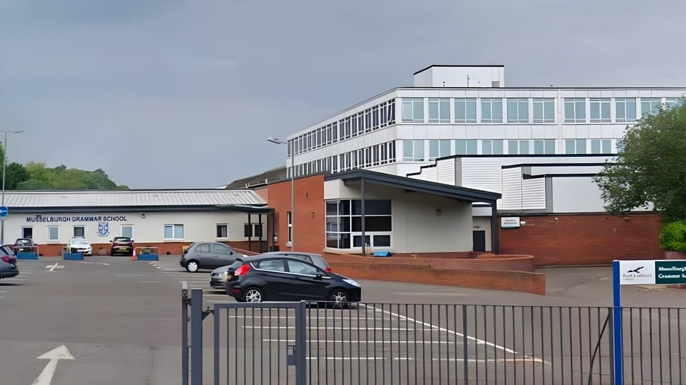 Das mehrstöckige Gebäude der Musselburgh Grammar School mit Glasfassade und Parkplatz im Vordergrund ist von Bäumen umgeben.