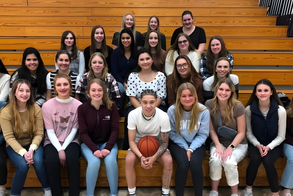 Schülerinnen der N.E.W. Lutheran High School sitzen gemeinsam auf Holzbänken in der Sporthalle mit einem Basketball im Vordergrund.