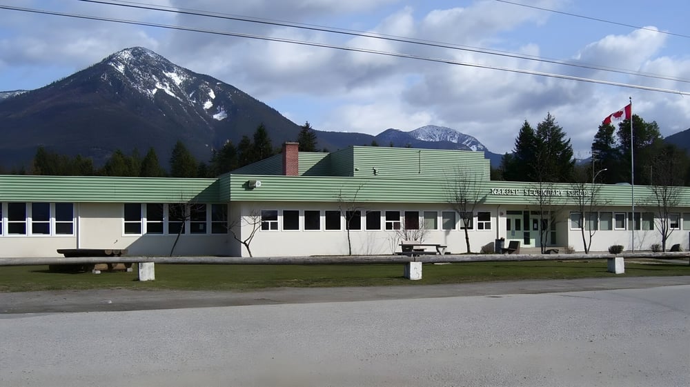 Das moderne mehrstöckige Gebäude der Nakusp Secondary School mit grünem Dach vor Bergen und Bäumen.