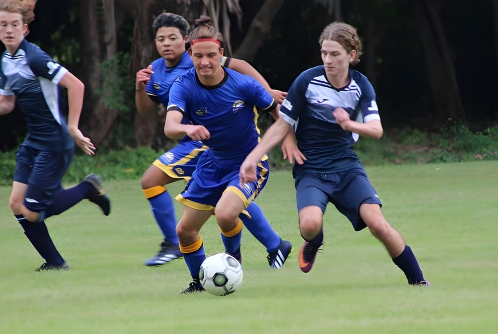 Schüler des Nambour State College spielen Fußball auf einem Rasenfeld mit Bäumen im Hintergrund.