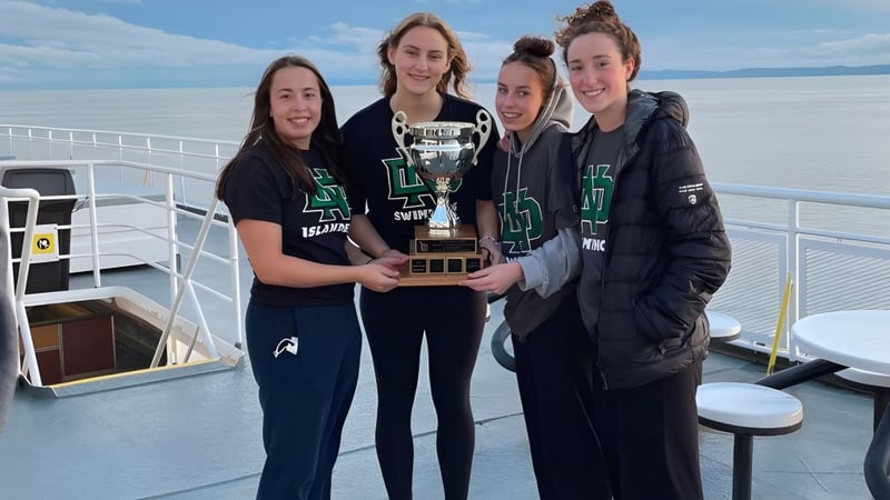 Eine Gruppe von vier Schülern der Nanaimo District Secondary School hält eine Trophäe auf dem Deck eines Bootes mit Wasser und bewölktem Himmel im Hintergrund.