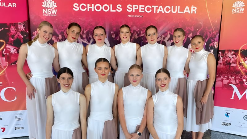 Schülerinnen der Narrabeen Sports High School posieren vor einem großen roten Banner mit der Aufschrift Schools Spectacular auf einer Bühne.