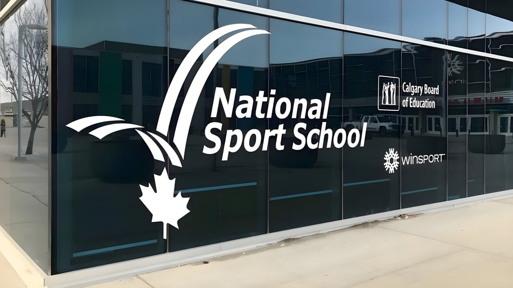 Das moderne Glasgebäude der National Sport School mit großem Schriftzug und Ahornblatt-Logo ist von außen zu sehen.