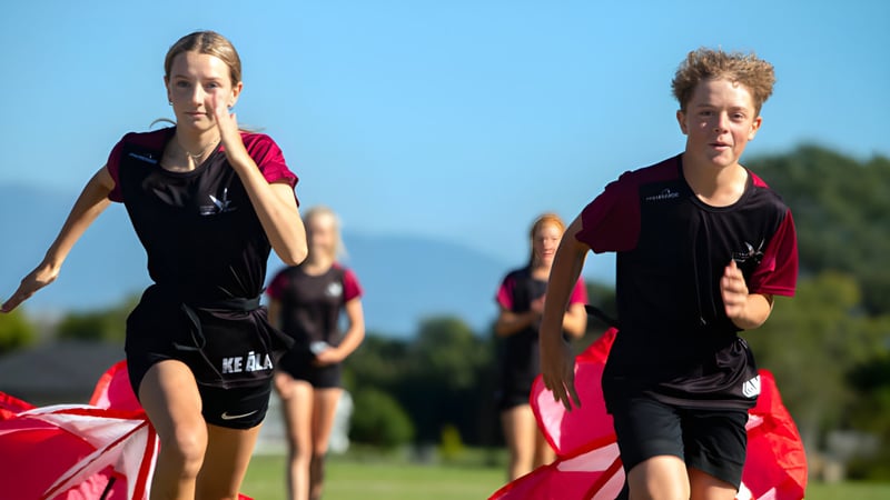 Zwei junge Athletinnen und Athleten laufen auf einem Rasenfeld vor Bergkulisse auf dem Campus des Nayland College.