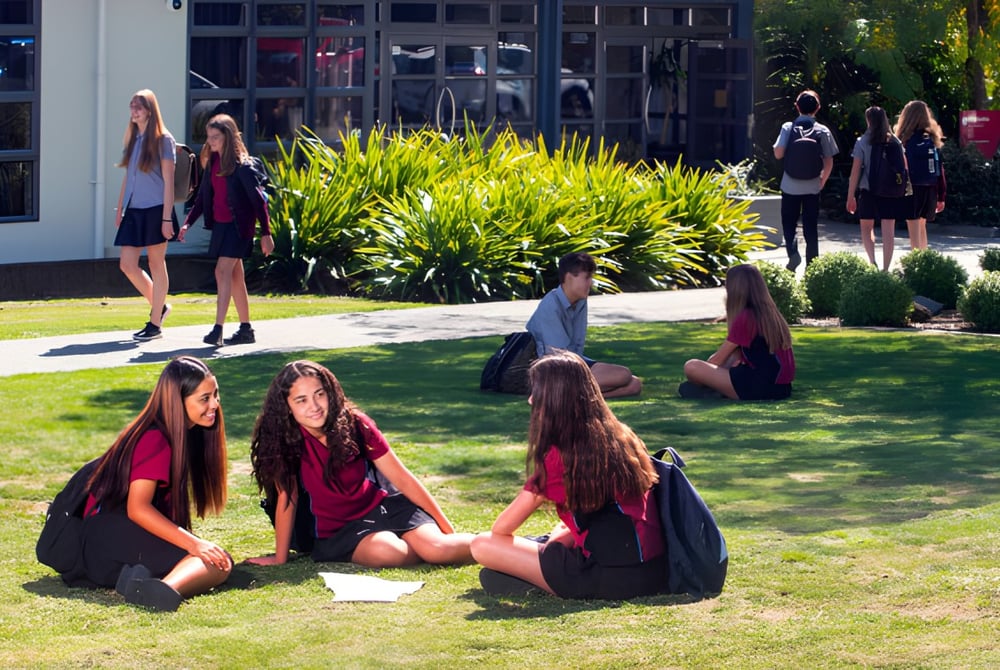 Eine Gruppe junger Frauen in schwarzen Talaren sitzt auf dem Gras vor einem Gebäude am Nayland College.