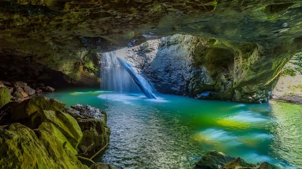 Ein türkisfarbener Wasserfall fließt über moosbedeckte Felsen in einen stillen Teich auf dem Gelände der Nerang State High School.