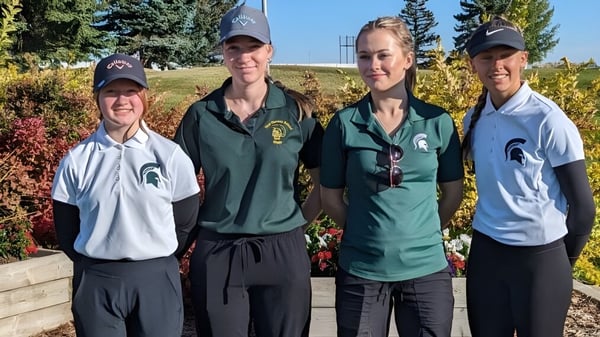 Vier Schülerinnen und Schüler der New Norway School in Michigan State University Sportbekleidung stehen vor einem bewaldeten Hintergrund unter klarem blauen Himmel.