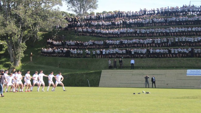 Schüler der New Plymouth Boys' High School stehen in weißen Uniformen auf einem Rasenfeld vor Zuschauern auf Tribünen.