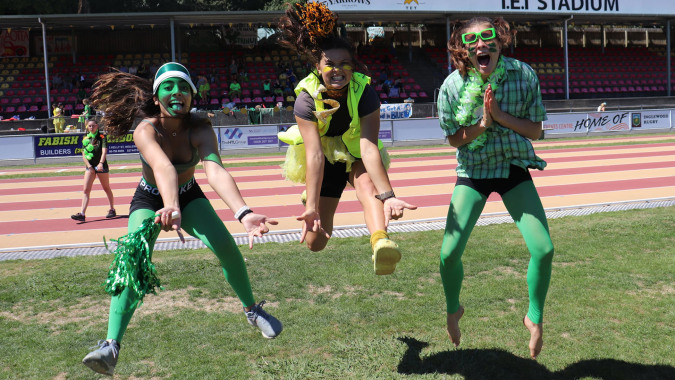Eine Gruppe von Schülerinnen der New Plymouth Girls’ High School trägt grüne Kostüme und versammelt sich auf einem Sportfeld mit Stadion im Hintergrund.