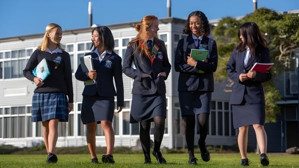 Fünf Schülerinnen in Uniformen stehen gemeinsam auf dem Campus der New Plymouth Girls’ High School mit einem Gebäude im Hintergrund.