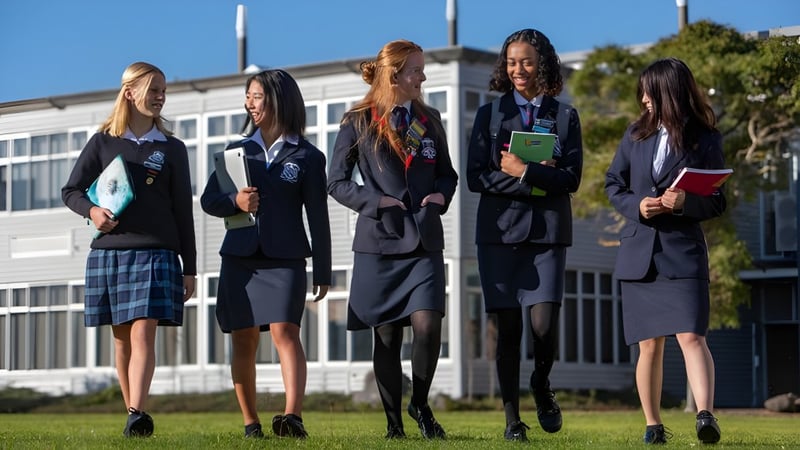 Fünf Schülerinnen in Uniformen stehen gemeinsam auf dem Campus der New Plymouth Girls’ High School mit einem Gebäude im Hintergrund.
