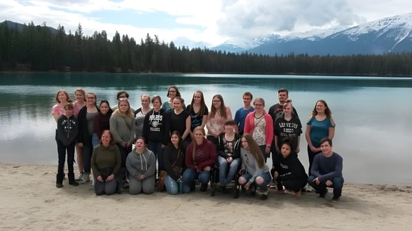 Schüler der New Sarepta Community High School versammeln sich an einem sandigen Strand vor einem See mit schneebedeckten Bergen im Hintergrund.