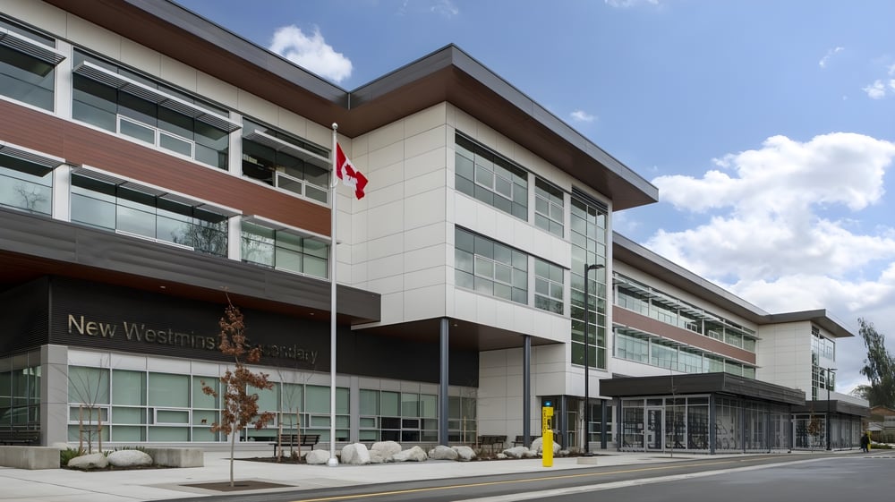 Das moderne mehrstöckige Gebäude der New Westminster Secondary School mit Glasfassade und gepflastertem Außenbereich ist bei bewölktem Himmel zu sehen.