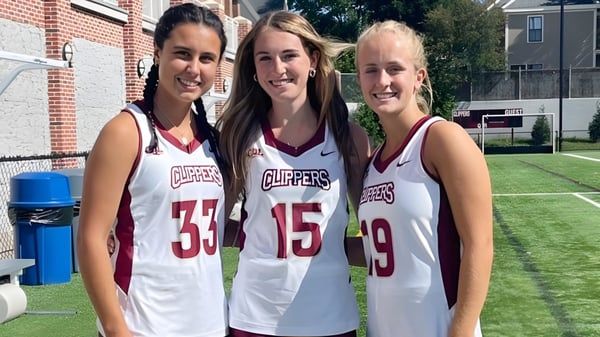 Drei Schülerinnen der Newburyport High School stehen im Sportjersey des Teams Clippers auf einem Rasen vor einem Backsteingebäude.
