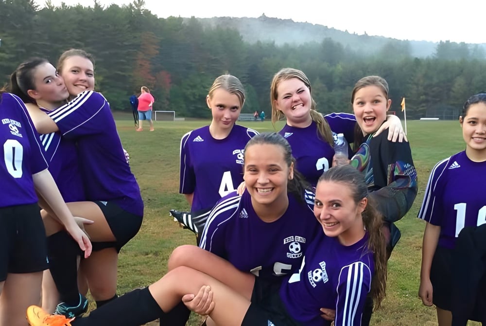 Eine Gruppe weiblicher Fußballspielerinnen des Newcomb Central School District steht auf dem Rasen eines Spielfelds mit Wald im Hintergrund.