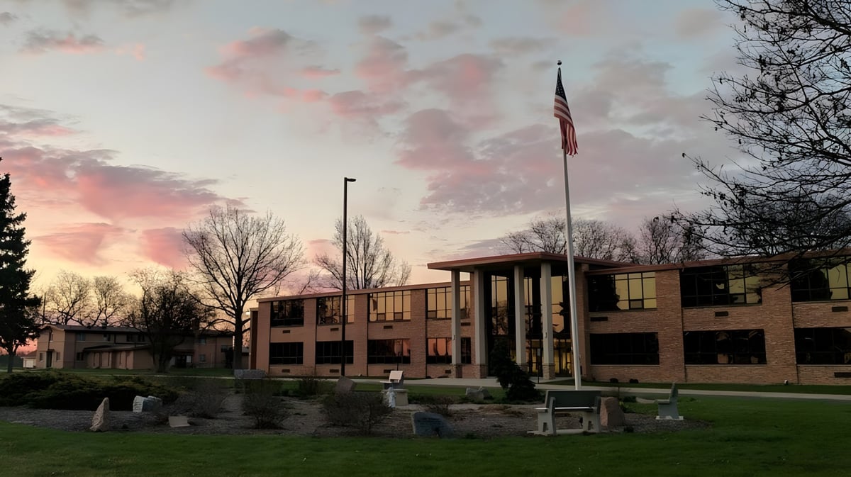 Das mehrstöckige Backsteingebäude der Newman Central Catholic High School vor einem bunten Sonnenuntergang mit Bäumen im Vordergrund.