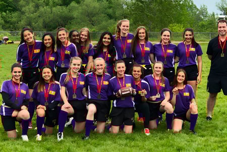 Eine Gruppe junger Fußballspielerinnen der Newmarket High School posiert gemeinsam auf einem Grasfeld mit Trainer im Hintergrund.