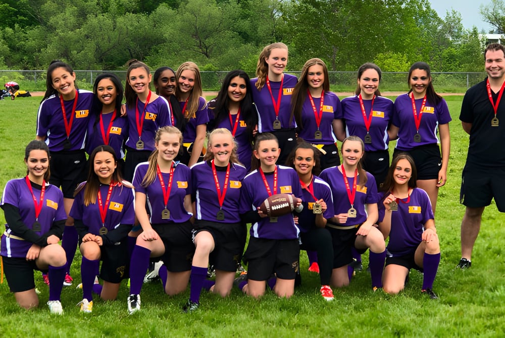 Eine Gruppe junger Fußballspielerinnen der Newmarket High School posiert gemeinsam auf einem Grasfeld mit Trainer im Hintergrund.
