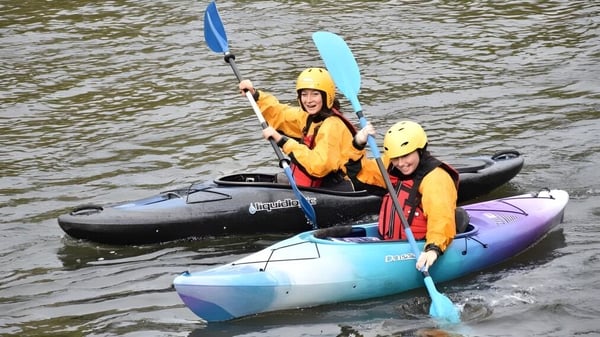 Eine Gruppe von Studierenden des Newstead College paddelt mit bunten Kajaks auf einem Fluss entlang eines bewaldeten Uferbereichs.