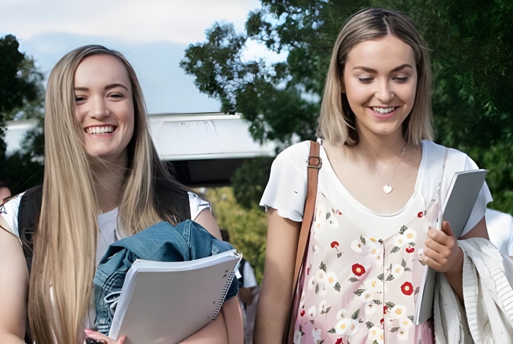 Zwei Schülerinnen des Newstead College gehen lächelnd über den Campus und tragen Bücher und Ordner.